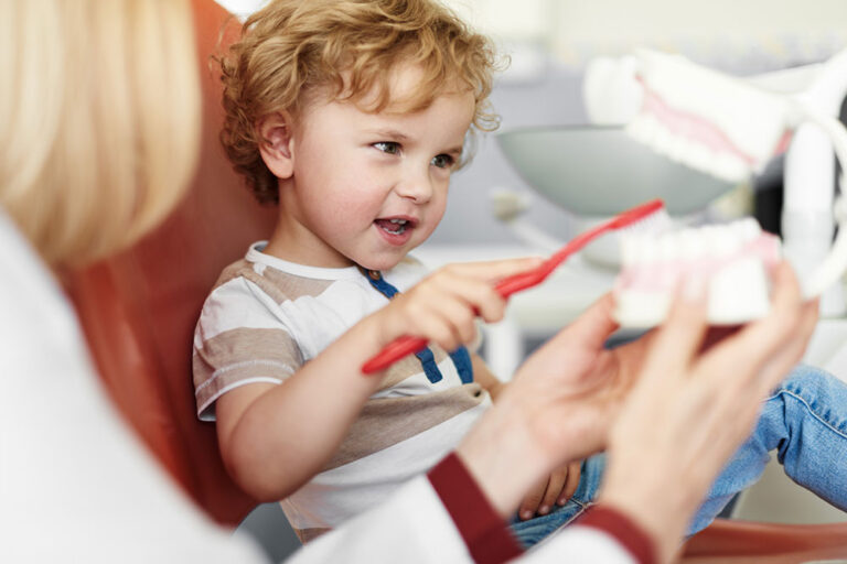 A young child seated in a dental chair, smiling while holding a red toothbrush and practicing on a dental model guided by a dental professional. The setting highlights a welcoming, child-friendly clinic focused on teaching proper oral hygiene.