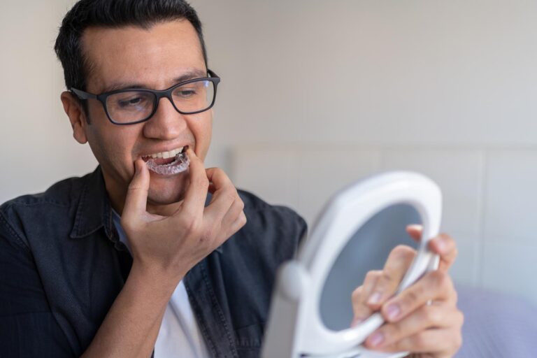 A man wearing glasses carefully inserts a clear dental night guard using a small mirror for guidance, demonstrating a precise orthodontic routine.