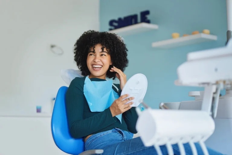 A patient sits in a dental chair wearing a bib and holding a round mirror to examine their teeth, surrounded by dental equipment in a clinic—capturing oral care and highlighting awareness of dental emergencies such as tooth pain, cracked teeth, or urgent treatment needs.